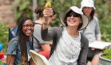 A woman holds up a squash blossom flower, smiling. Several educators are in the background, seated in a garden outdoors.