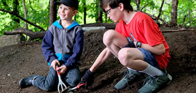 Two young students dig in school garden with forest in background. One student smiles off camera.