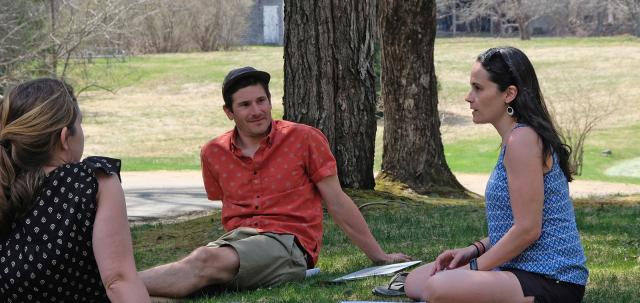 Three educators sit in grass talking in early spring. They look engaged and focused.