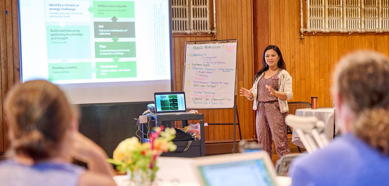 An educator stands at front of room in Shelburne Farms' Coach Barn, next to an easel and projector screen, conducting a presentation. In foreground, two educators sit at a table with backs to camera.